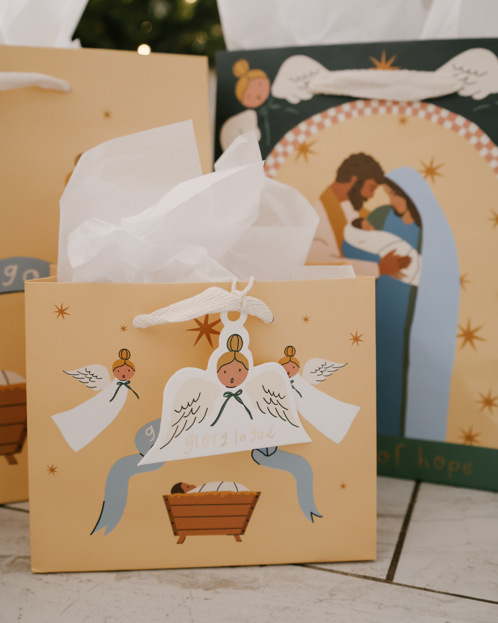 Christmas gift bag sets featuring Nativity scenes with baby Jesus, Mary, and Joseph, angels, and starry skies. Includes 5 bags in 3 sizes per set, with cotton ribbon handles and matching gift tags. Designs include 'Glory to God' and 'A Thrill of Hope.'