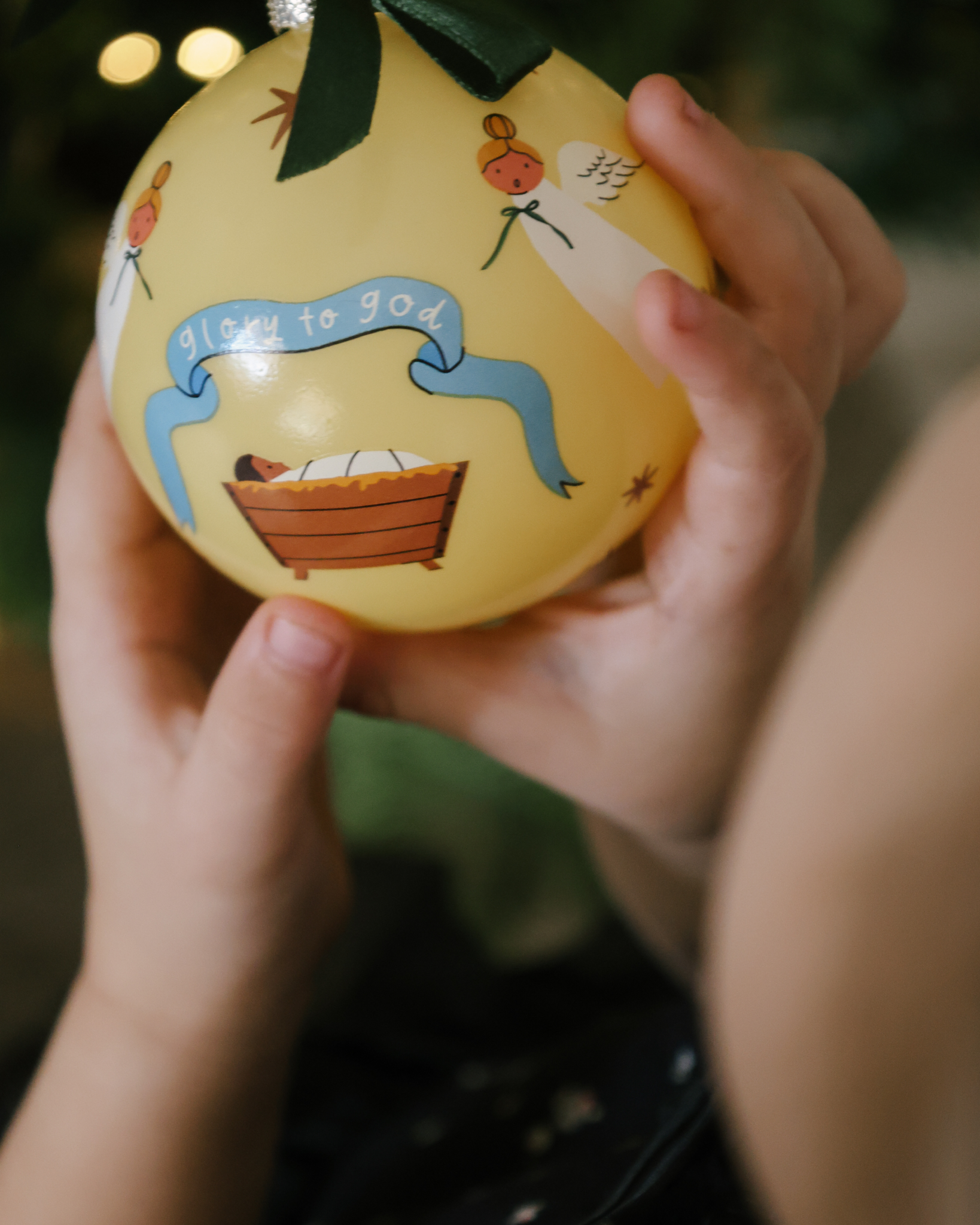 Glory to God Christmas ornament showing baby Jesus in the manger surrounded by angels, with satin ribbon hanger. Made of shatterproof plastic, ideal for families.