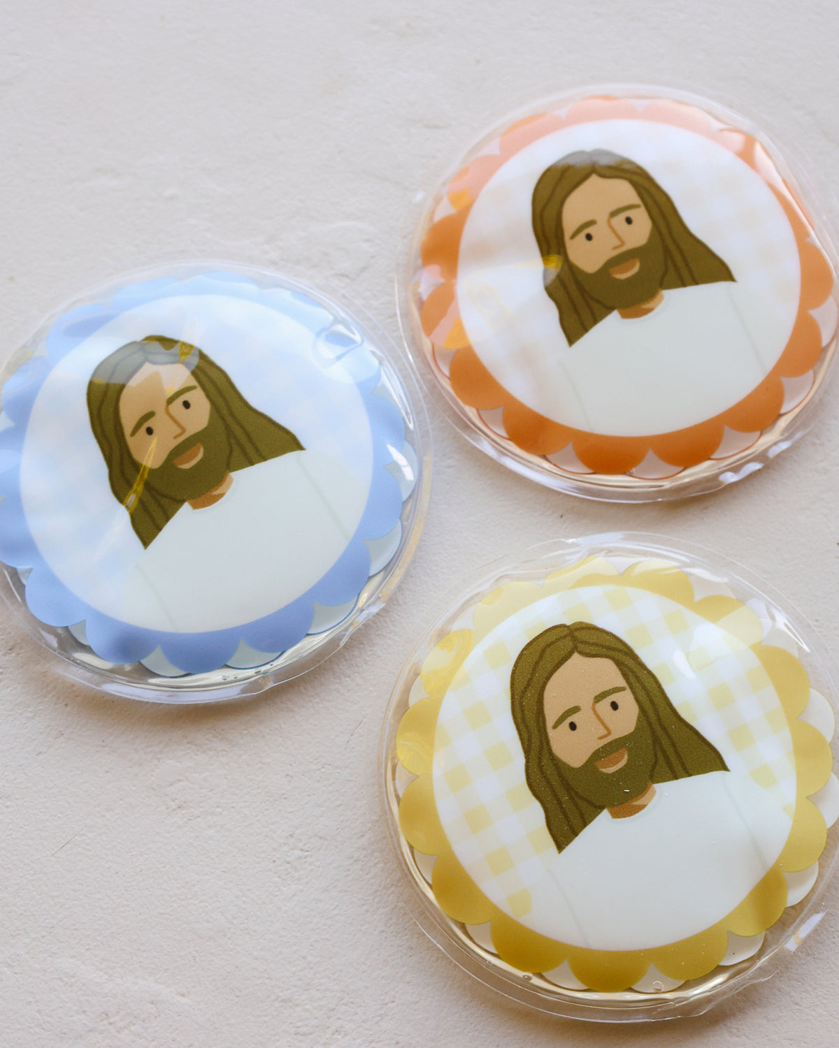 Close up of Jesus Heals hot and cold gel packs with scalloped gingham borders in blue, orange and yellow